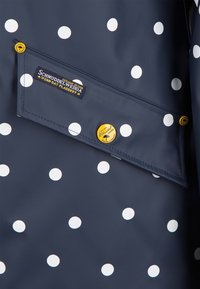 Tissu bleu marine à pois blancs, avec un rabat en tissu avec une étiquette et trois boutons jaunes avec du texte noir et des icônes de parapluie.
