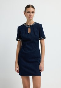 Femme portant une robe bleu marine à manches courtes avec un col orné de bijoux, des poignets de manches ornés de bijoux et un décolleté en trou de serrure, se tenant devant un fond blanc.