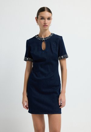 Vrouw in een donkerblauwe jurk met korte mouwen, voorzien van een juweelrijke kraag, juweelrijke mouwmanchetten en een keyhole halslijn, staande tegen een witte achtergrond.
