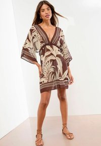 Kaftan marrone e crema con motivo, scollatura a V profonda, maniche ampie, vita cinchata e orlo a strati. Indossato con sandali marroni con cinturini.
