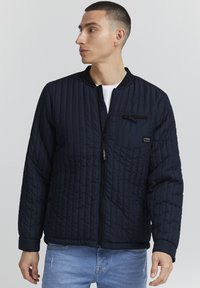 Jachetă din material navy, matlasată, cu buzunar cu fermoar, guler și manșete cu dungi. Prezintă un design texturat și o închidere cu fermoar pe întreaga față.