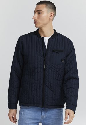 Marineblå quiltet jakke med glidelåslomme, ribbestrikket krage og mansjetter. Har et strukturert design og en full front glidelås.