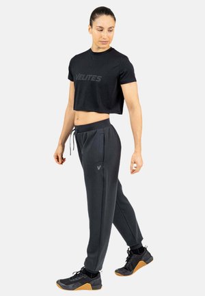Mujer con top corto negro con texto "VELITES", pantalones deportivos gris oscuro con logo "V" y zapatillas deportivas negras con suela de goma, de pie y mirando hacia abajo.