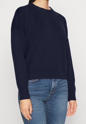Mørkeblå sweater med rund halsudskæring, ribbede manchetter og kant, lavet af et blødt, tekstureret stof. Kombineret med tætsiddende denimjeans.