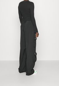 Zwarte geribbelde langemouwen top en losse zwarte broek met ruches en gespsluitingen. Draagt witte sneakers met groene accenten.