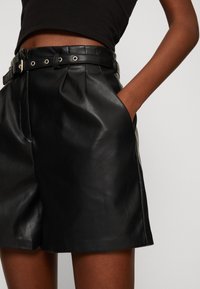 Shorts en cuir noir taille haute avec une large ceinture comportant des œillets en métal, deux poches latérales et une texture lisse.