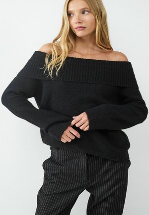 Schwarzer, off-the-shoulder Strickpullover mit geripptem Halsausschnitt und lockerer Passform. Kombiniert mit schwarzen Nadelstreifenhosen. Weicher Stoff, lässiges Design.