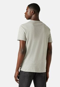 Hombre de pie de espaldas, vistiendo una camiseta de manga corta gris claro y jeans gris oscuro, contra un fondo liso.