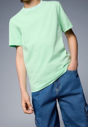 Osoba ubrana w jasnozieloną koszulkę Calvin Klein Jeans i niebieskie dżinsy z jedną ręką w kieszeni na szarym tle.