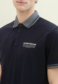 Mornarska polo majica z teksturiranim sivočrnim ovratnikom, tremi gumbi in belimi logotipnimi detajli. Kratki rokavi s črtastimi poudarki na manšetah.