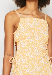 Žluté květinové šaty s bílými a růžov�ými vzory, nastavitelné boční stahování a hranatý výstřih. Tkanina má strukturovaný povrch.