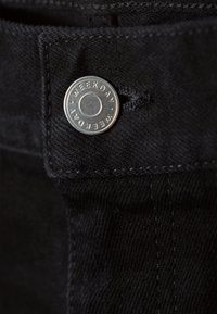 Bouton argenté avec le mot "WEEKDAY" embossé sur un tissu en denim noir près d'une boutonnière cousue.