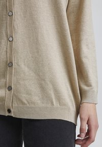 Cardigan lavorato a maglia beige con chiusura a bottoni metallici, polsini e orlo a costine e una texture morbida. Indossato sopra jeans scuri.