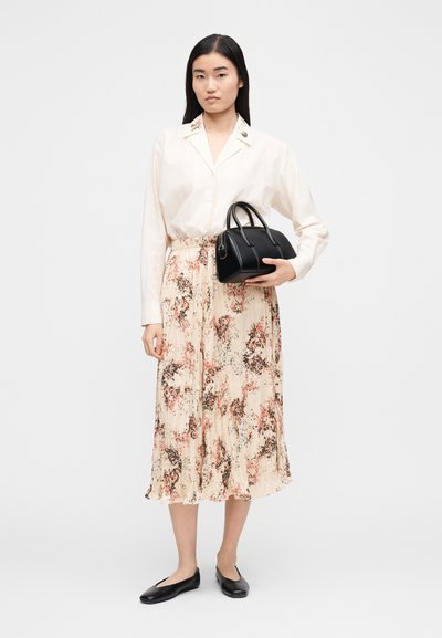 Woman wearing a cream blouse with floral collar and a pleated floral skirt, holding a black handbag and wearing black flats on white background.
