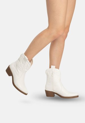 Ben iført hvide læderankelstøvler med lave brune hæle, med sidelommer og syede dekorative mønstre, mod en ensartet baggrund.