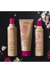 Trois produits capillaires Aveda Cherry Almond : un shampoing et un soin sans rinçage dans des flacons cylindriques dorés avec des bouchons roses, et un après-shampoing en tube doré avec un motif floral.