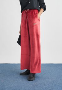 Osoba stojící na modrém koberci, oblečená v červených sametových kalhotách s širokými nohavicemi a stahovací šňůrkou v pase, v černých teniskách, s rukou v kapse.