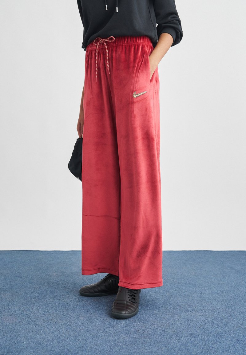 Osoba stojící na modrém koberci, oblečená v červených sametových kalhotách s širokými nohavicemi a stahovací šňůrkou v pase, v černých teniskách, s rukou v kapse.