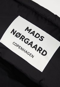 Czarny materiał z białą metką, na której widnieje napis "MADS NØRGAARD COPENHAGEN" w wyraźnym, pogrubionym czarnym tekście. Tekst jest czytelny i wyraźnie umiejscowiony.