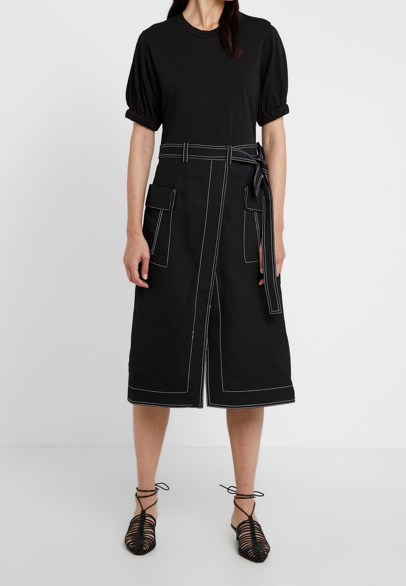 Femme portant un haut noir à manches bouffantes et une jupe noire taille haute avec des coutures blanches, une fente frontale, de grandes poches et une ceinture nouée.