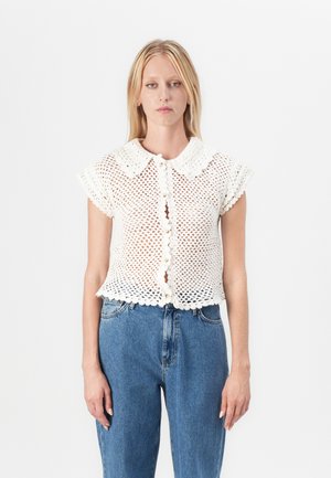 White knit short-sleeve top with a scalloped collar, button closure, and an open weave design. Paired with high-waisted blue denim jeans.