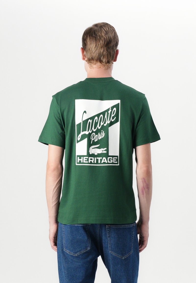 T-shirt en coton vert avec un grand imprimé graphique blanc au dos, affichant "Lacoste Paris" et "HERITAGE", accompagné d'un logo crocodile.