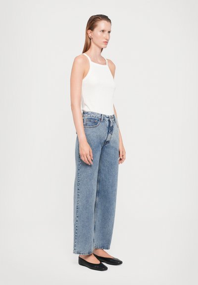 White sleeveless top paired with high-waisted, wide-leg blue denim jeans. Model wears black flats; smooth texture with classic pocket details.