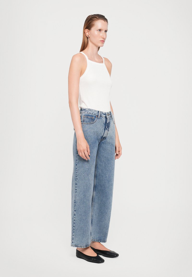 Haut sans manches blanc associé à un jean bleu en denim à taille haute et jambe large. Le mannequin porte des ballerines noires ; texture lisse avec des détails de poches classiques.