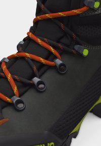 Κοντινό πλάνο μαύρου πέδιλου ορειβασίας με μαύρα και πορτοκαλί κορδόνια που περνάνε μέσα από μεταλλικά τελάρα και πράσινες λεπτομέρειες στη σόλα και το πάνω μέρος.