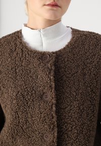 Cardigan en peluche marron à texture shaggy avec un col rond, doté de trois boutons marron. Porté sur un haut blanc à col montant en matière côtelée.