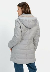 Grå polstret jakke med hætte, der har en blød pelsforing, vandret quilting og en lidt længere pasform. Båret over blå jeans.