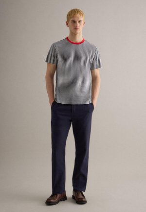 Jonge man staand met handen in de zakken, gekleed in een zwart-wit gestreept T-shirt met rode kraag, marineblauwe broek en bruine schoenen, tegen een effen achtergrond.