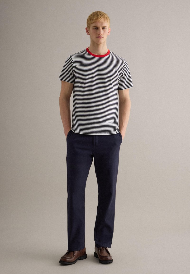 Giovane in piedi con le mani nelle tasche, indossa una maglietta a righe bianco e nere con colletto rosso, pantaloni blu navy e scarpe marroni su uno sfondo neutro.