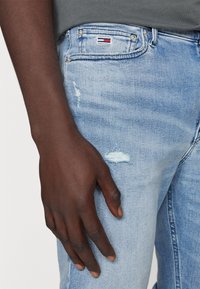 Jeans de mezclilla de color azul claro con detalles desgastados, incluyendo parches deshilachados. Presenta una etiqueta con el logo en el bolsillo delantero. Mano reposando sobre el muslo.
