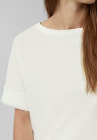 Biała bawełniana koszulka z krótkimi, podwiniętymi rękawami, okrągłym dekoltem, miękką fakturą i minimalistycznym wzornictwem, prezentująca gładkie wykończenie.