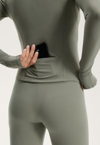 Rukávová, priliehavá blúzka v olivovo zelenej farbe s vreckom na zips na zadnej strane, v ktorom sa nachádza čierny smartfón, s malým detailom loga.