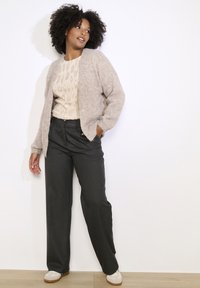 Gai bēšs kardigāns ar pūkainu tekstūru, atvērta priekšpuse. Bēšs rakstains džemperis zemā. Tumši pelēkas platajās bikses, krēmīgas krāsas sporta apavi. Ikdienišķs apģērbs.