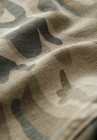 Kamuflaažkangas, millel on tekstureeritud pind, koos beezide, roheliste ja tumedate pruunide toonidega. Abstraktne muster kõverate kujundite ja joontega.