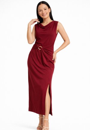 Kvinde poserende i ærmeløs bordeaux kjole med sideslids og guldringdetalje, iført beige ankelstroppesko.