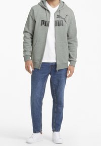 Felpa grigia con zip e logo nero, abbinata a una t-shirt bianca a scollo a V e jeans in denim blu. Sneakers bianche completano l'outfit.