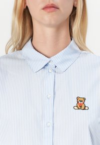 Blå- och vitrandig skjorta med knäppkrage och en liten broderad nallebjörn på det vänstra bröstområdet. Slät tygtextur.