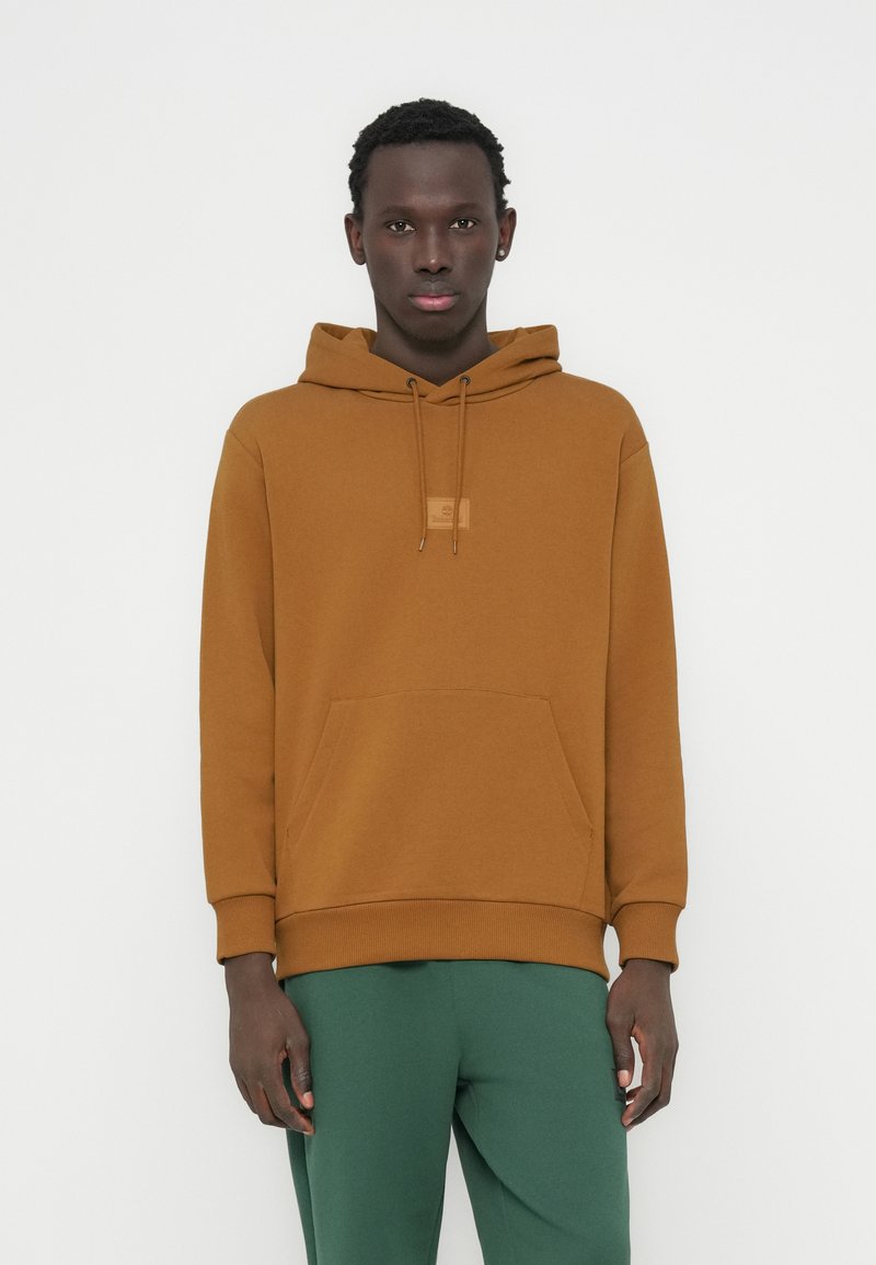 Καφέ φούτερ από βαμβάκι με τσέπη καγκουρό, κουκούλα με ρυθμιζόμενο κορδόνι και μανσέτες με ρίγες, συνδυασμένο με πράσινη φόρμα.