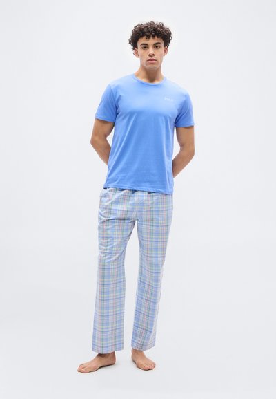 Νεαρός άνδρας που στέκεται ξυπόλητος φορώντας μπλε μπλουζάκι πόλο και ανοιχτόχρωμο καρό παντελόνι πιτζάμας σε απλό φόντο.