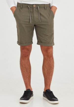 Shorts vert olive en tissu léger, avec un cordon de serrage aux accents blancs, des ourlets retroussés et des poches latérales. Portés avec des baskets noires.