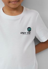 Witte t-shirt met korte mouwen en een ronde halslijn. Bevat een logo in zwarte en teal tekst op de linkerborst: "ALLEEN DE WILD DUNE WINDEN WETEN."