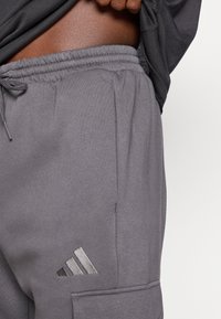 Pantalones de chándal grises con cintura elástica, textura suave, bolsillo lateral y un logotipo reflectante negro de Adidas en el muslo izquierdo.