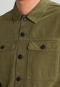 Veste chemise vert olive en tissu durable, dotée d'un col classique, de deux poches avant et de boutons noirs pour la fermeture.