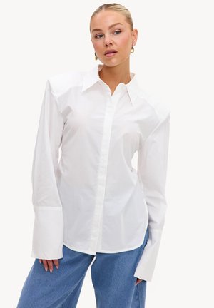Femme portant une chemise blanche oversize à boutons avec des poignets allongés et un jean bleu, regardant légèrement sur le côté, les cheveux tirés en arrière.