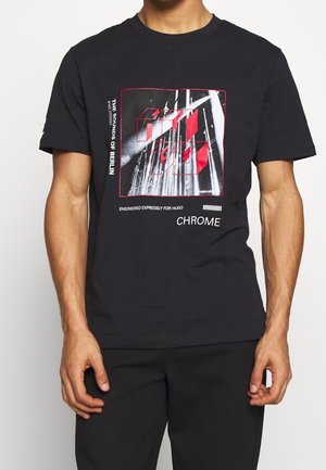 Muž v černém tričku s červeným a bílým abstraktním motivem, s nápisy "THE SOUNDS OF BERLIN," "ENGINEERED EXPRESSLY FOR HUGO" a "CHROME."