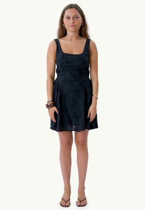Jeune femme aux longs cheveux portant une robe noire sans manches, des boucles d'oreilles, des bracelets et des tongs, debout devant un fond blanc uni.